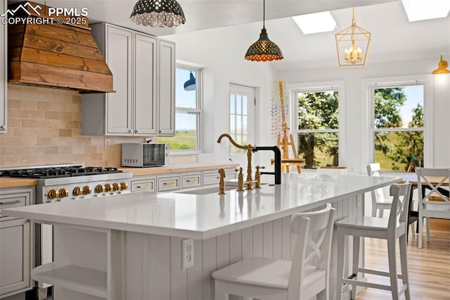 kitchen featuring sink, a kitchen island with sink, hanging light fixtures, custom range hood, and decorative backsplash