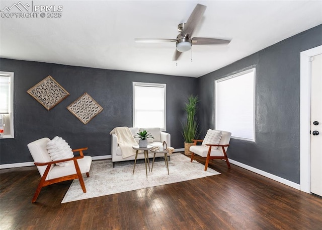 living area with an accent wall, dark wood finished floors, baseboards, and a ceiling fan