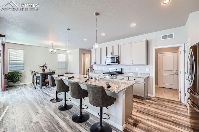 kitchen featuring pendant lighting, gray cabinets, stainless steel appliances, light stone counters, and an island with sink