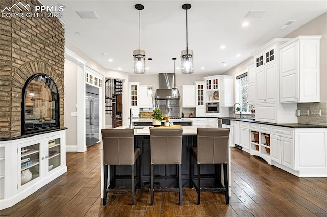 kitchen featuring decorative light fixtures, a center island with sink, appliances with stainless steel finishes, wall chimney range hood, and white cabinets