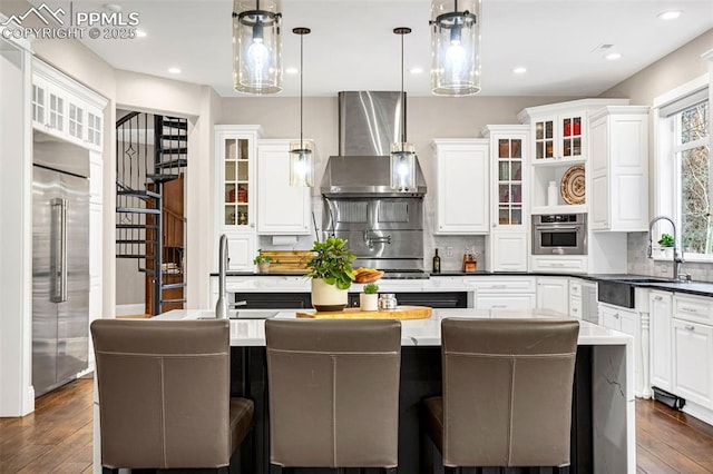 kitchen with wall chimney range hood, stainless steel appliances, a center island, white cabinets, and decorative light fixtures