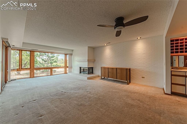 unfurnished living room with ceiling fan, a multi sided fireplace, a textured ceiling, brick wall, and light colored carpet