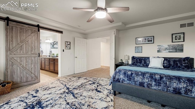 bedroom featuring ensuite bathroom, light colored carpet, a tray ceiling, ceiling fan, and a barn door