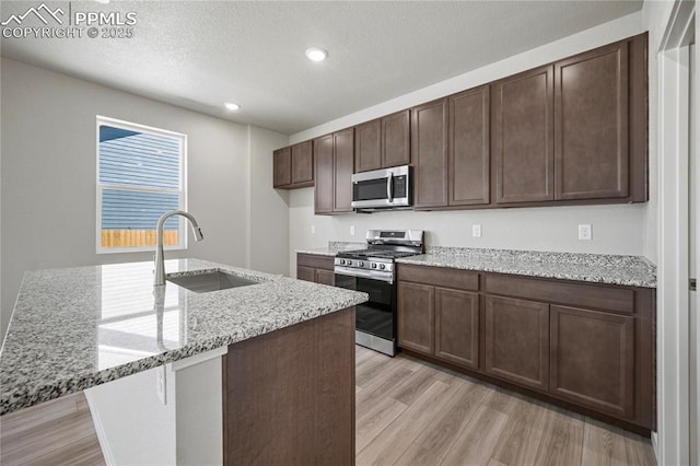 kitchen with sink, appliances with stainless steel finishes, dark brown cabinets, light stone countertops, and light hardwood / wood-style floors