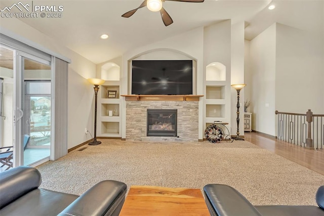 carpeted living room with vaulted ceiling, a stone fireplace, ceiling fan, and built in shelves