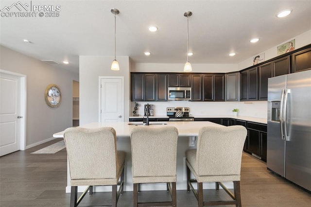 kitchen with dark hardwood / wood-style floors, appliances with stainless steel finishes, decorative light fixtures, and an island with sink