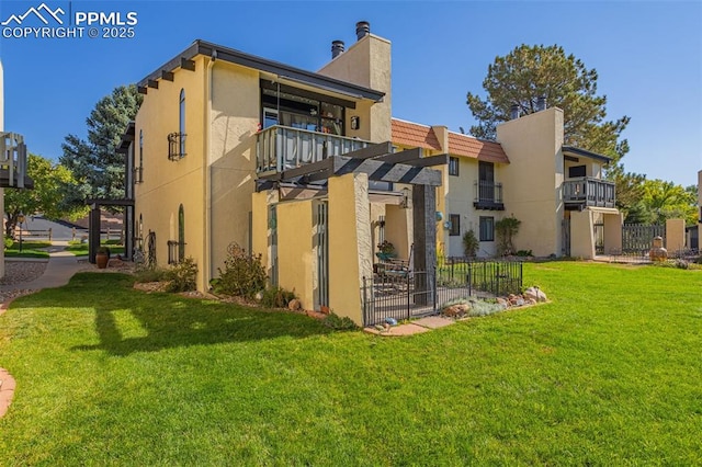 back of house featuring a lawn, a balcony, a chimney, fence, and stucco siding