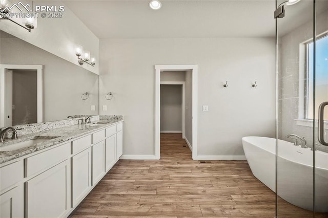 full bathroom with double vanity, a sink, wood finished floors, a freestanding tub, and baseboards