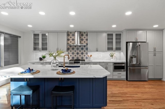 kitchen with wall chimney range hood, a breakfast bar area, appliances with stainless steel finishes, light stone counters, and a center island with sink