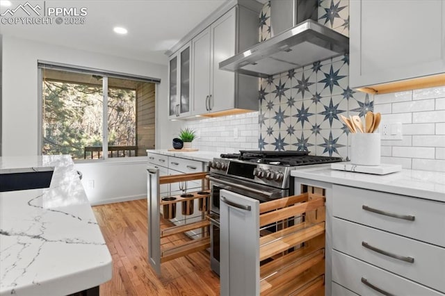 kitchen with wall chimney range hood, stainless steel gas range, backsplash, light stone counters, and light wood-type flooring