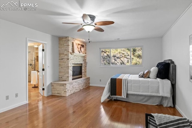 bedroom with a stone fireplace, ensuite bathroom, ceiling fan, and light wood-type flooring