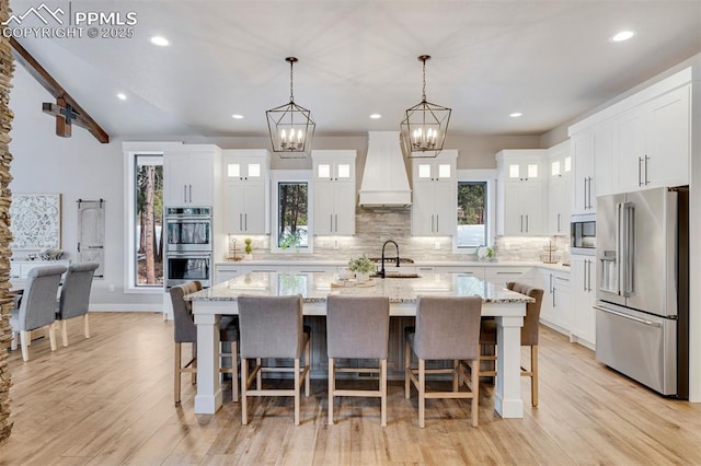 kitchen with custom exhaust hood, appliances with stainless steel finishes, a center island with sink, and white cabinets