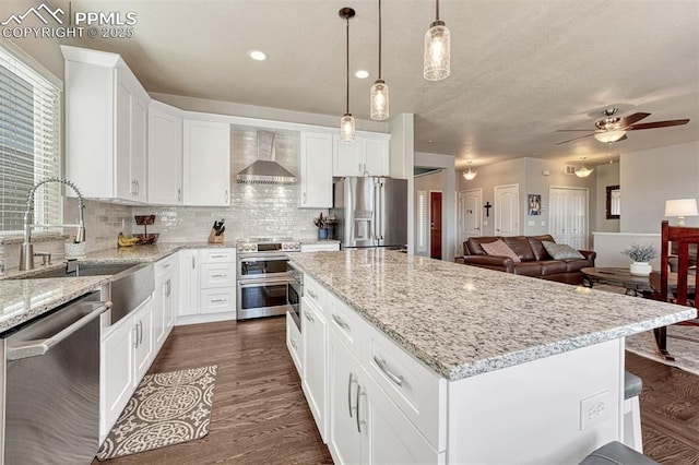 kitchen featuring a kitchen island, appliances with stainless steel finishes, white cabinetry, hanging light fixtures, and wall chimney exhaust hood