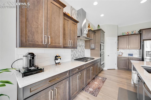 kitchen with appliances with stainless steel finishes, backsplash, light stone countertops, wall chimney exhaust hood, and light wood-type flooring