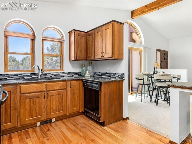 kitchen with dishwasher, brown cabinets, dark stone countertops, vaulted ceiling with beams, and a sink