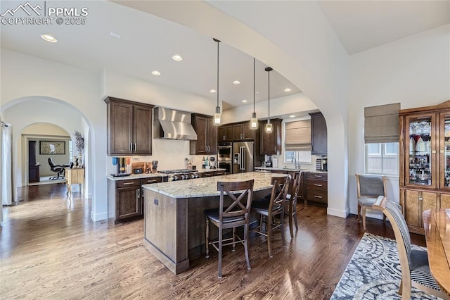 kitchen with pendant lighting, wall chimney range hood, dark brown cabinetry, light stone counters, and a center island with sink