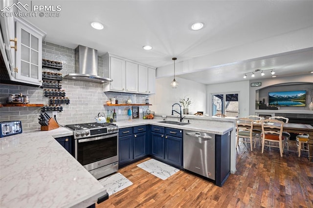 kitchen with wall chimney range hood, sink, stainless steel appliances, white cabinets, and blue cabinets