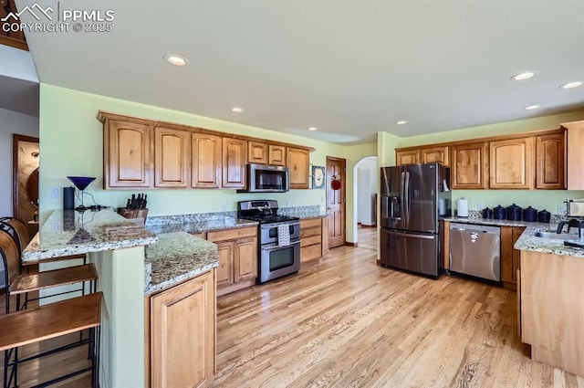 kitchen with arched walkways, light wood-style flooring, stainless steel appliances, a peninsula, and light stone countertops