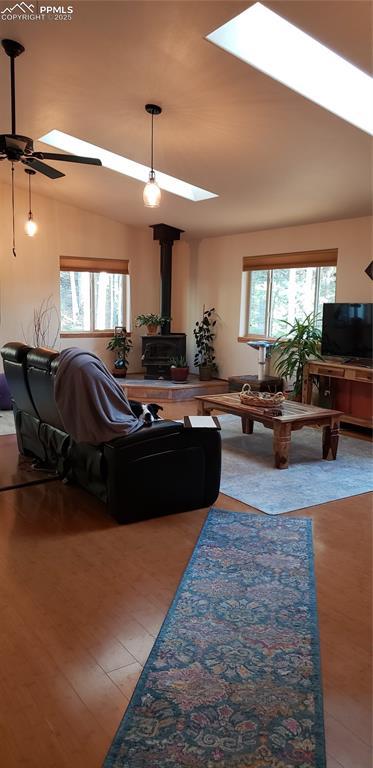 living area featuring a wood stove, lofted ceiling with skylight, light wood finished floors, and a ceiling fan