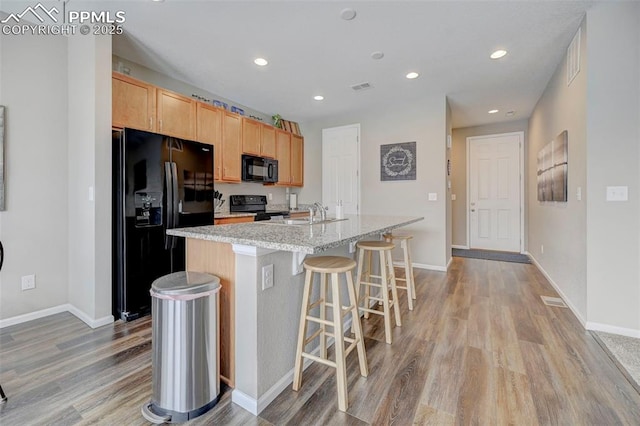 kitchen with a breakfast bar area, light stone counters, light hardwood / wood-style floors, black appliances, and an island with sink