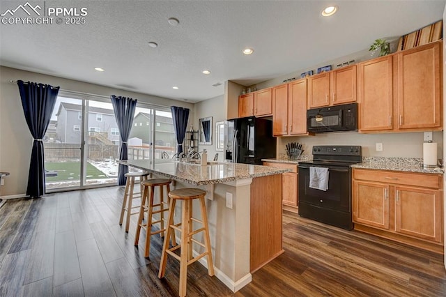 kitchen featuring a breakfast bar area, a kitchen island with sink, dark hardwood / wood-style floors, light stone counters, and black appliances