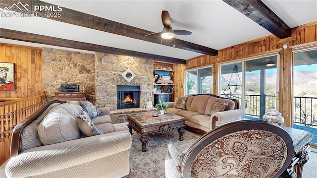 living room with ceiling fan, wooden walls, beamed ceiling, and a stone fireplace