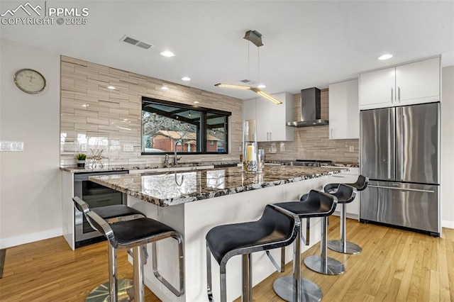 kitchen featuring wall chimney exhaust hood, dark stone counters, visible vents, white cabinetry, and stainless steel refrigerator