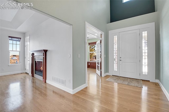 entrance foyer featuring light wood-style flooring, a towering ceiling, visible vents, baseboards, and a glass covered fireplace