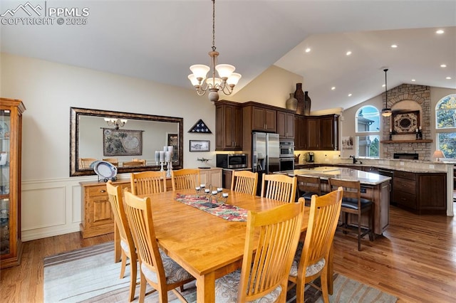 dining room featuring a decorative wall, light wood-style floors, wainscoting, vaulted ceiling, and ceiling fan with notable chandelier