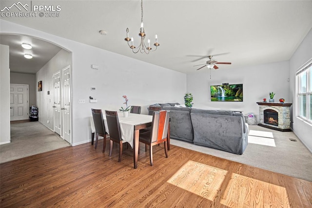 dining room with arched walkways, a stone fireplace, ceiling fan with notable chandelier, wood finished floors, and baseboards