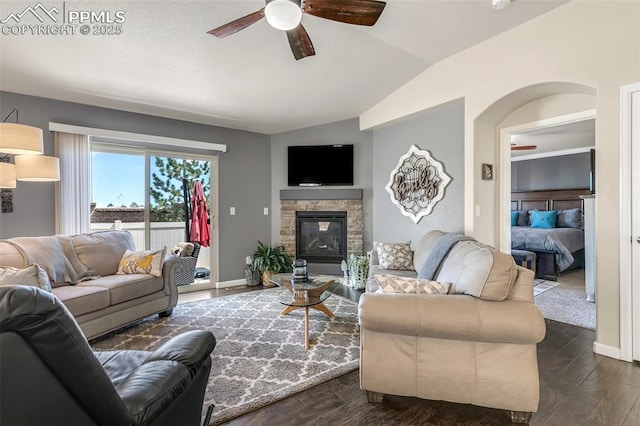 living area featuring lofted ceiling, dark wood-type flooring, ceiling fan, a stone fireplace, and baseboards