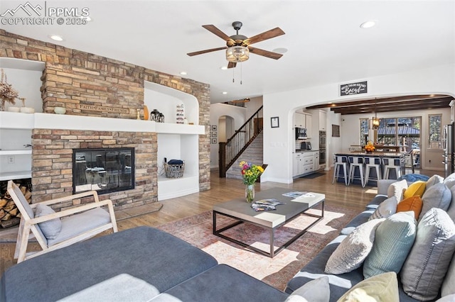 living area with arched walkways, ceiling fan, wood finished floors, stairs, and a fireplace