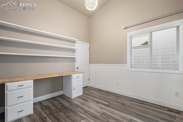 unfurnished office with dark wood-style flooring, built in desk, visible vents, wainscoting, and vaulted ceiling