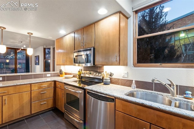 kitchen with dark tile patterned flooring, a sink, appliances with stainless steel finishes, brown cabinetry, and pendant lighting