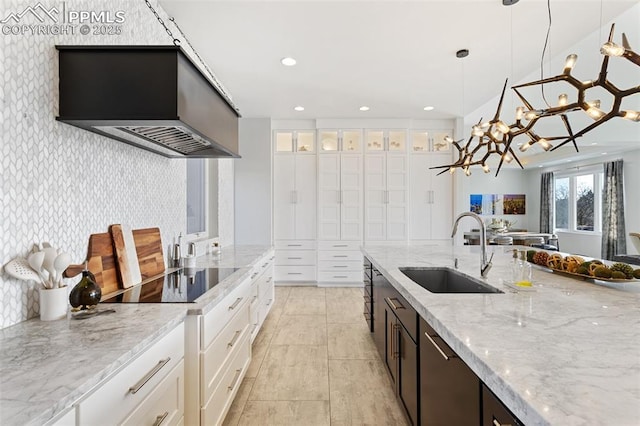 kitchen with custom range hood, hanging light fixtures, light stone countertops, white cabinetry, and a sink