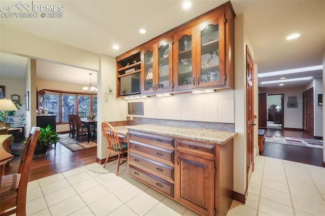 kitchen with a chandelier, light stone counters, light tile patterned flooring, decorative backsplash, and glass insert cabinets