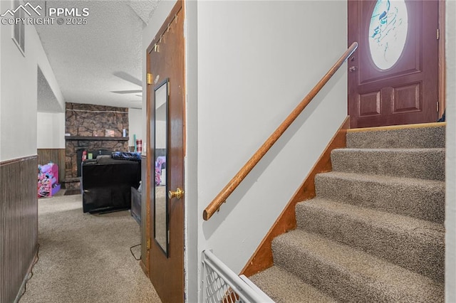 stairway featuring wainscoting, carpet, a textured ceiling, a stone fireplace, and wood walls