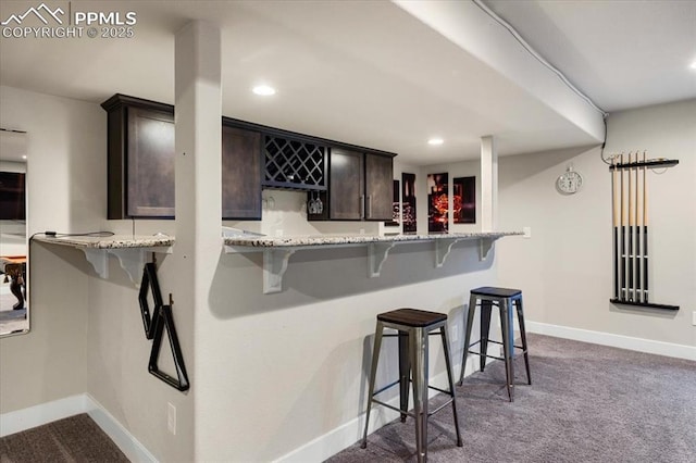 kitchen featuring light stone counters, dark brown cabinetry, baseboards, dark carpet, and a kitchen bar