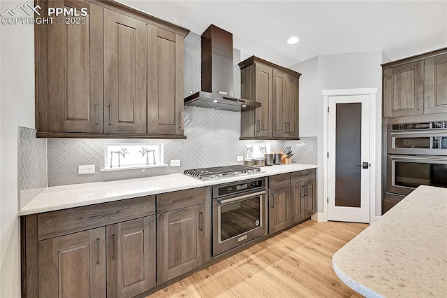 kitchen with stainless steel appliances, light wood-style floors, light stone countertops, wall chimney exhaust hood, and tasteful backsplash