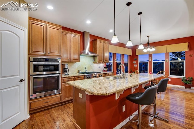 kitchen with stainless steel appliances, hanging light fixtures, a sink, wall chimney range hood, and an island with sink