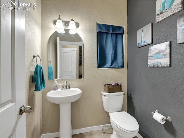 half bath with baseboards, a textured wall, toilet, tile patterned flooring, and a sink