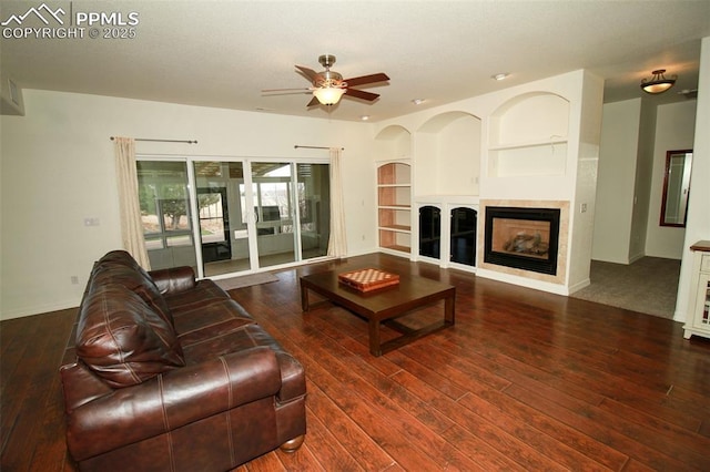 living area featuring baseboards, wood-type flooring, ceiling fan, built in shelves, and a fireplace