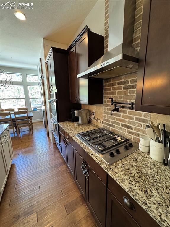 kitchen with tasteful backsplash, appliances with stainless steel finishes, dark brown cabinetry, wood finished floors, and wall chimney exhaust hood