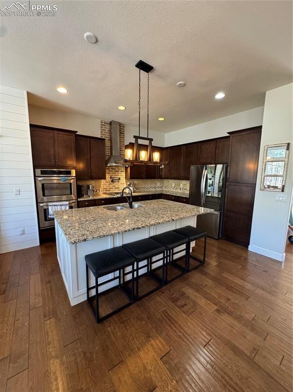 kitchen with a sink, appliances with stainless steel finishes, wall chimney exhaust hood, an island with sink, and pendant lighting