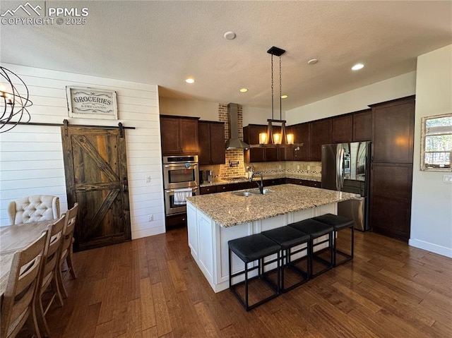 kitchen with stainless steel appliances, a barn door, a kitchen island with sink, a sink, and light stone countertops