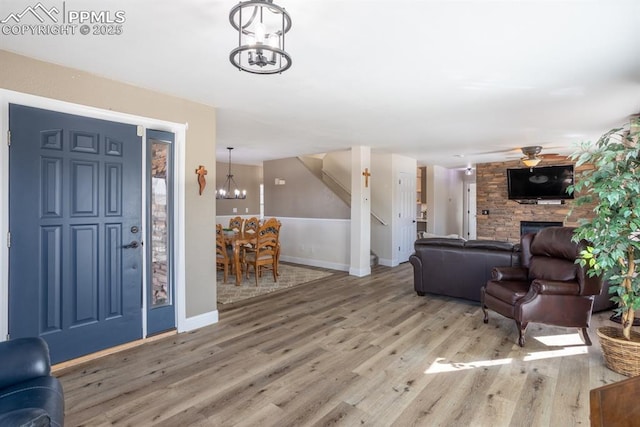 foyer entrance with stairs, a stone fireplace, light wood-type flooring, baseboards, and ceiling fan with notable chandelier