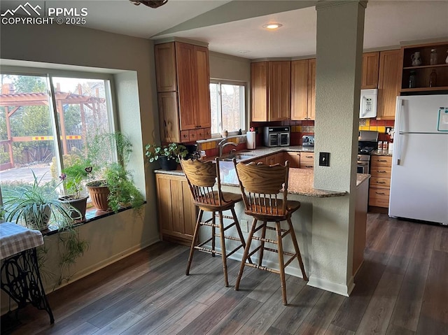 kitchen with a peninsula, white appliances, brown cabinetry, and dark wood-type flooring