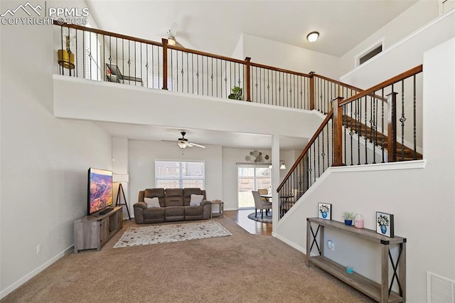 carpeted living area with visible vents, a towering ceiling, a ceiling fan, baseboards, and stairs