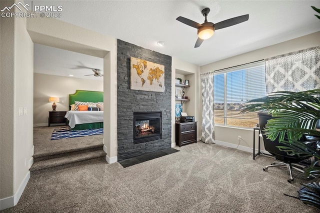 carpeted office space featuring a stone fireplace, built in shelves, a ceiling fan, and baseboards