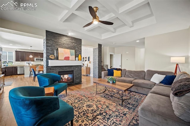 living area featuring beam ceiling, coffered ceiling, a stone fireplace, and light wood-style flooring